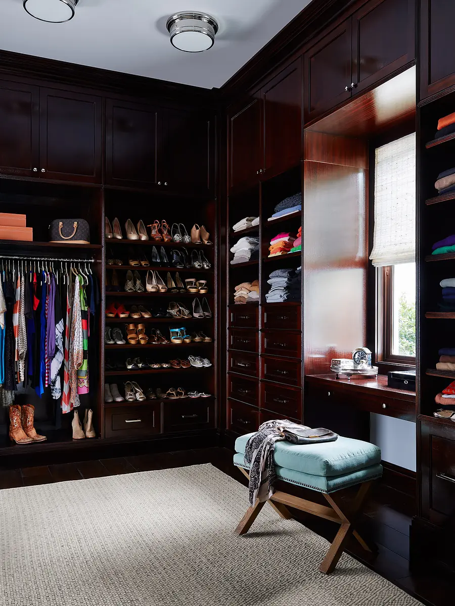 Closet with dark wood shelves, shoes, clothing, window, rug, and upholstered stool.