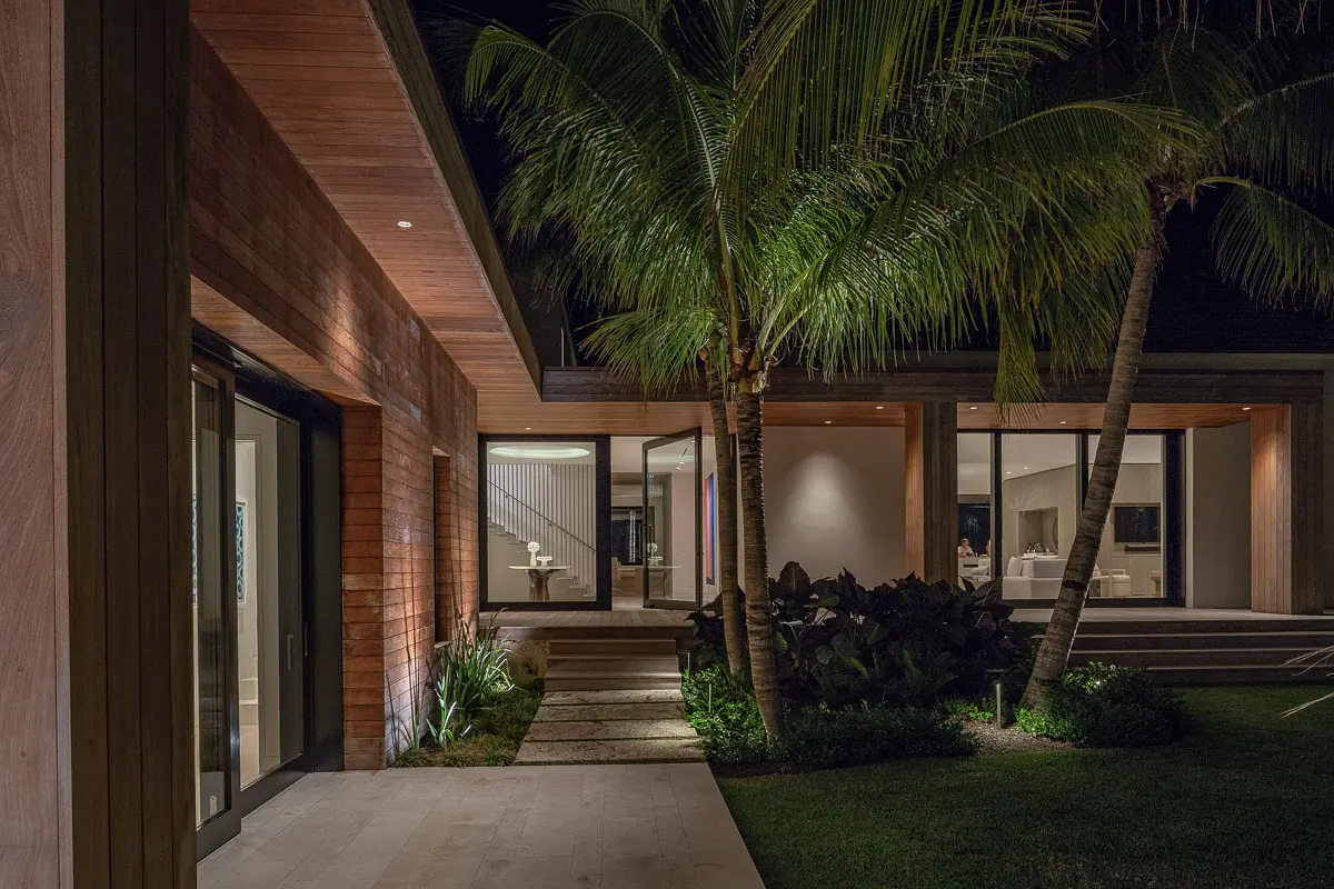 Exterior front view of a house with wood cladding, large glass doors, palm trees, and stone pathways.