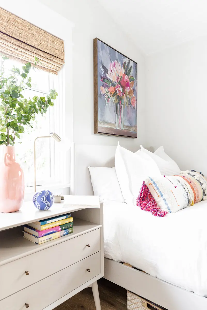 Bedroom with bed, nightstand, pink vase, blue decor, desk lamp, wall art, and woven shade.