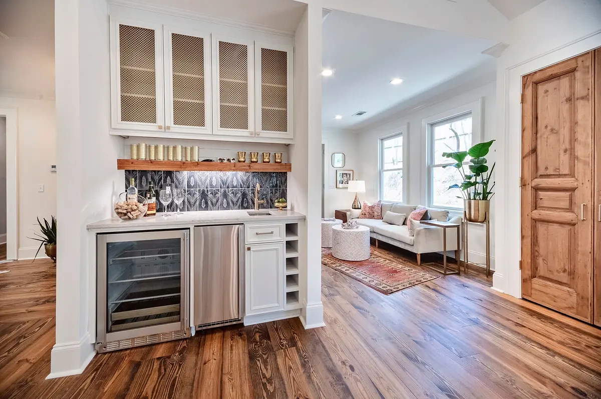 Butler's pantry with wine cooler, wood cabinetry, backsplash, and adjacent living area with sofa and rug.