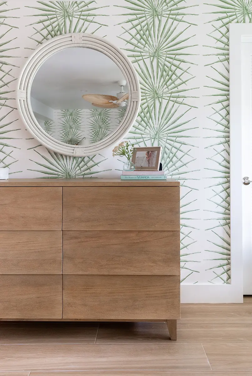 Bedroom with wooden dresser, round mirror, palm leaf wallpaper, and light-colored tiled floor