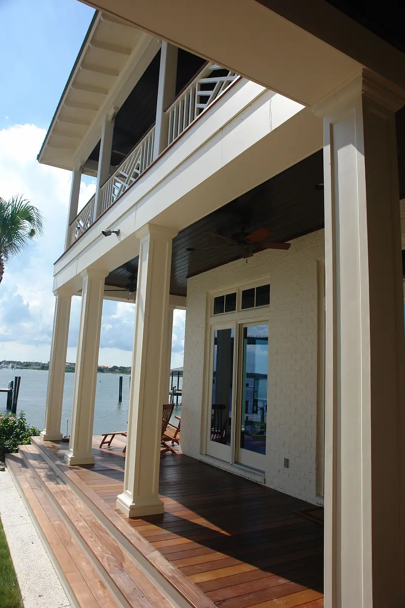 Outdoor deck with wooden flooring, large columns, railing, ceiling fan, and rocking chairs overlooking water.