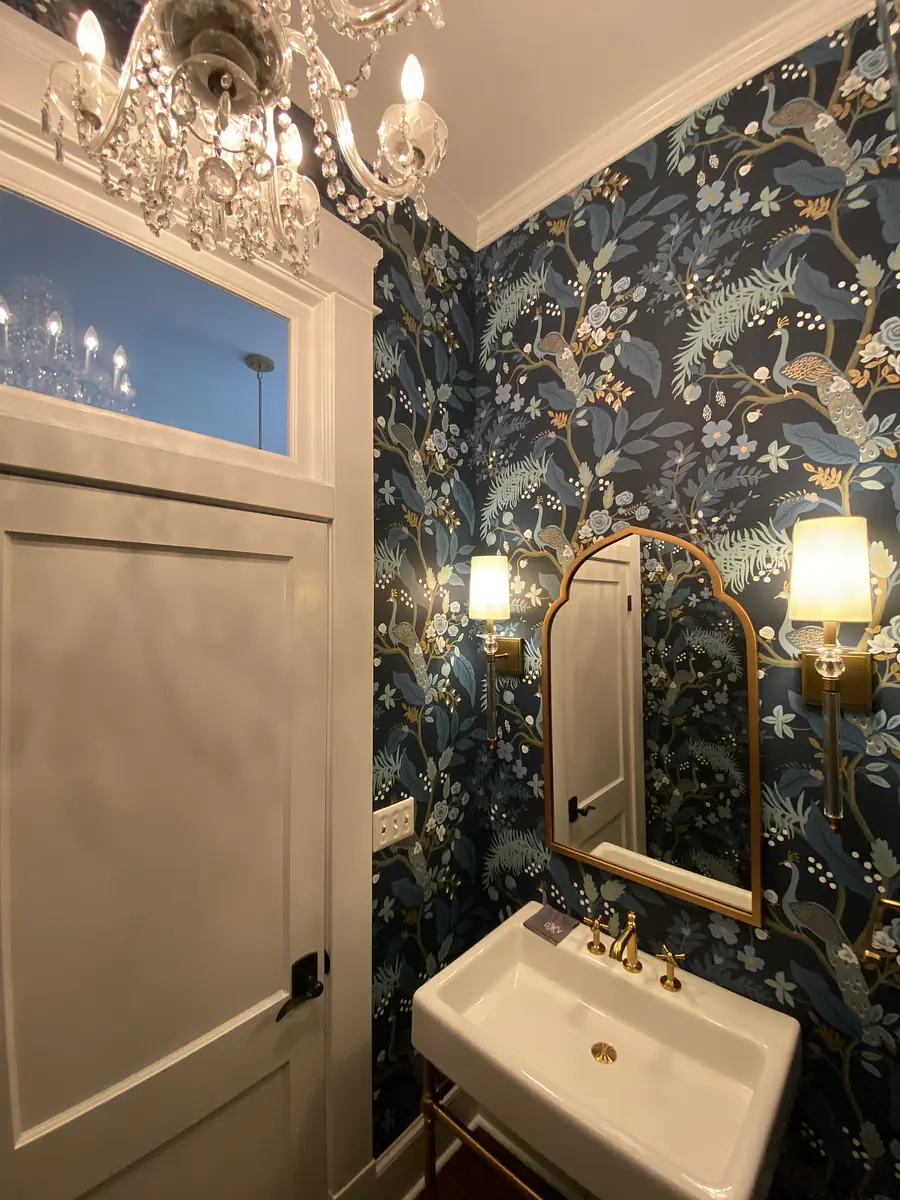Powder room with floral wallpaper, white pedestal sink, gold fixtures, and wall-mounted mirror.