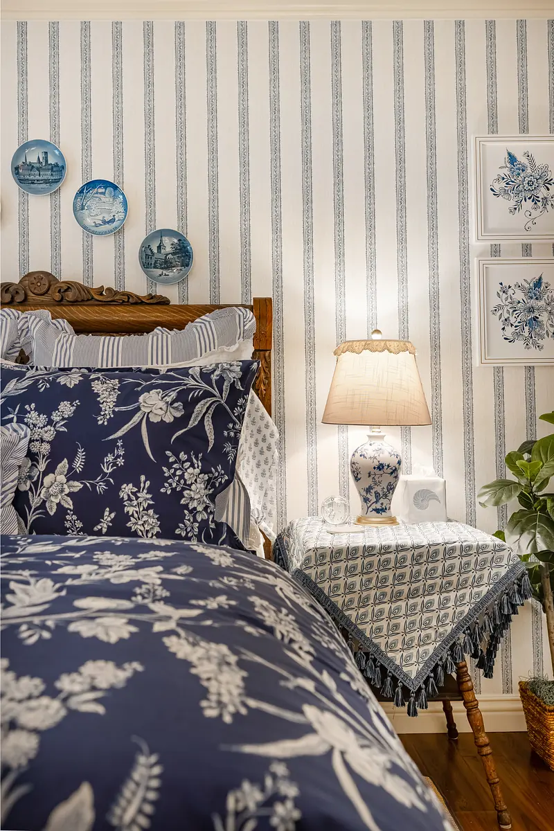 Bedroom with wooden bed, navy bedding, nightstand with lamp, striped wallpaper, and blue decorative plates.