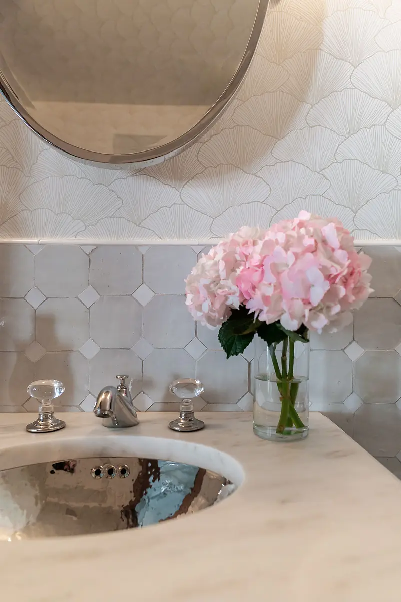 Bathroom with marble countertop, circular mirror, chrome faucet, and pink hydrangeas in a glass vase.