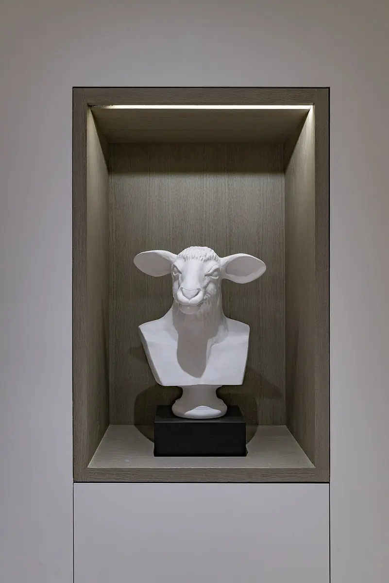 Display niche with white sheep bust on black base against wood panel and overhead lighting.