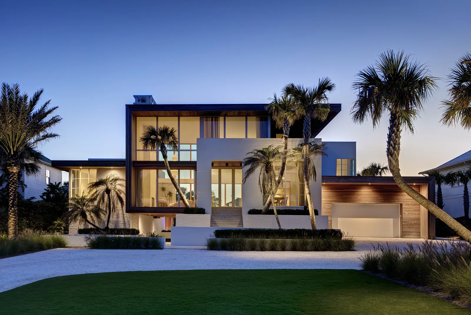 Front exterior view of a house with large windows, wooden accents, palm trees, and a spacious driveway.