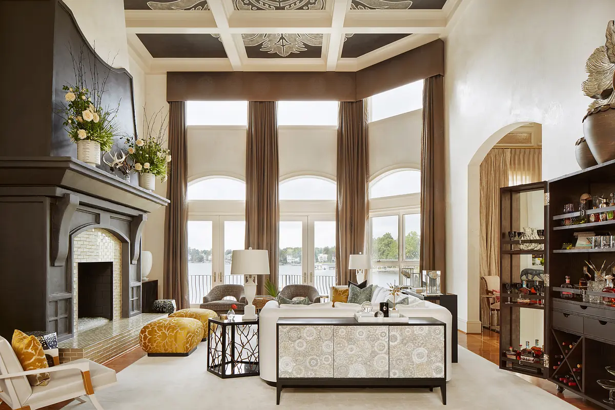 Living room with large fireplace, beige sofa, patterned ottoman, glass coffee table, and tall windows overlooking water.