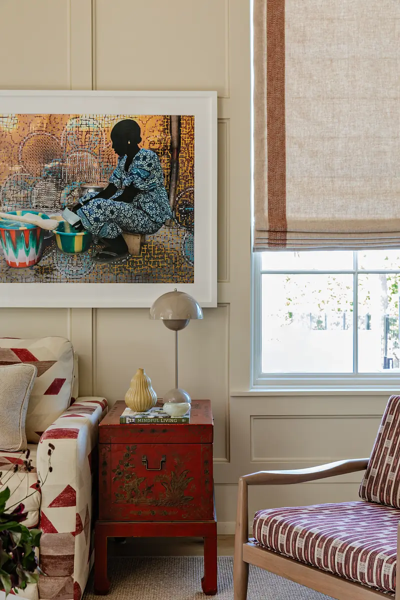 Living room with patterned sofa, striped side chair, red cabinet, artwork, and window with roll-up shade.