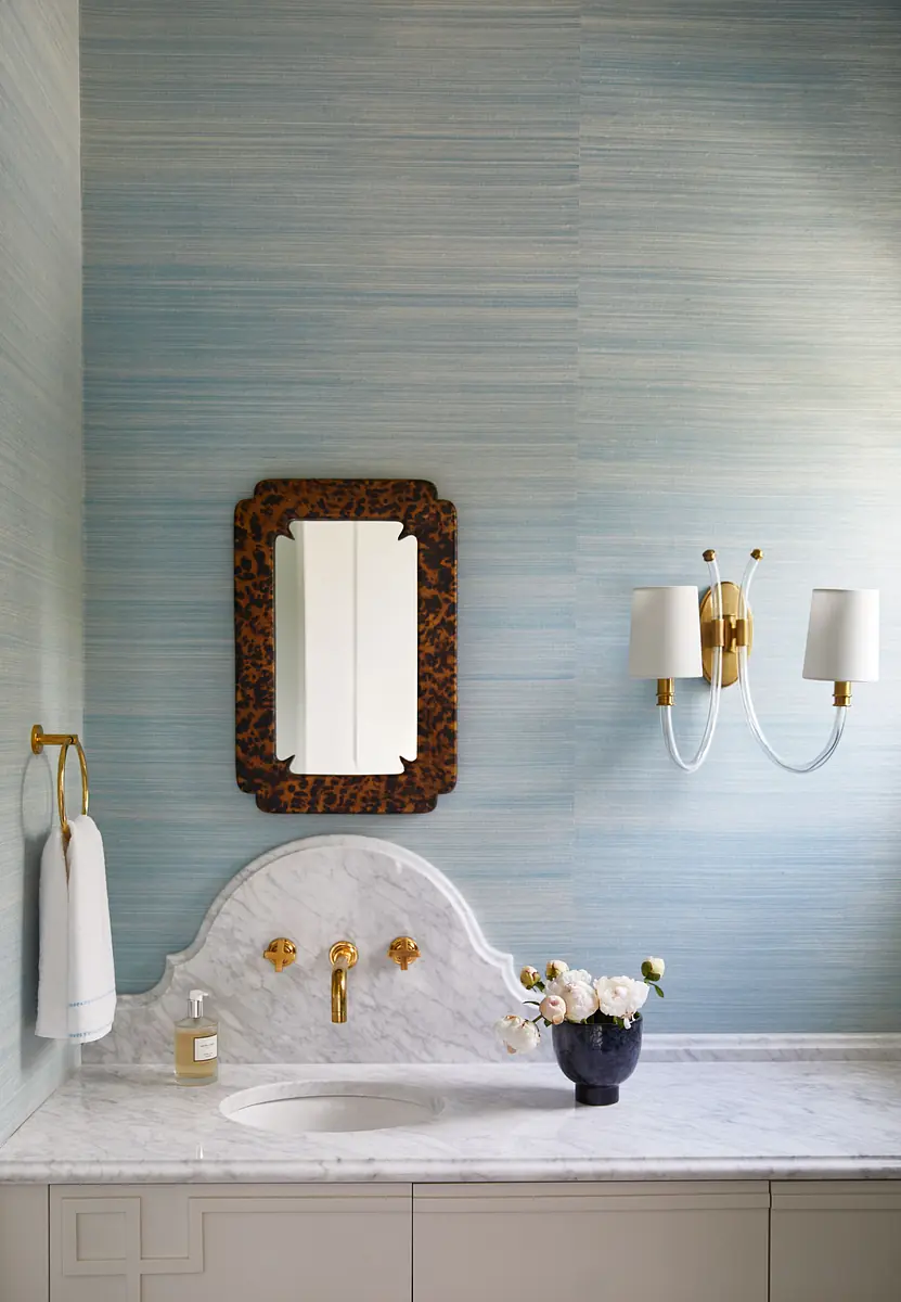 Bathroom with marble countertop, wall-mounted mirror, gold sconce, and blue striped wallpaper.