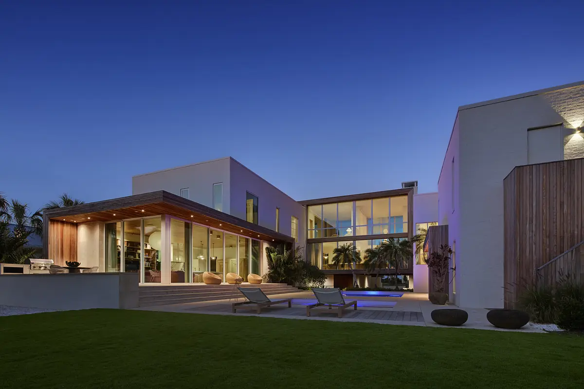 Exterior rear view of a house with large glass windows, wooden deck, lounge chairs, and landscaped lawn.