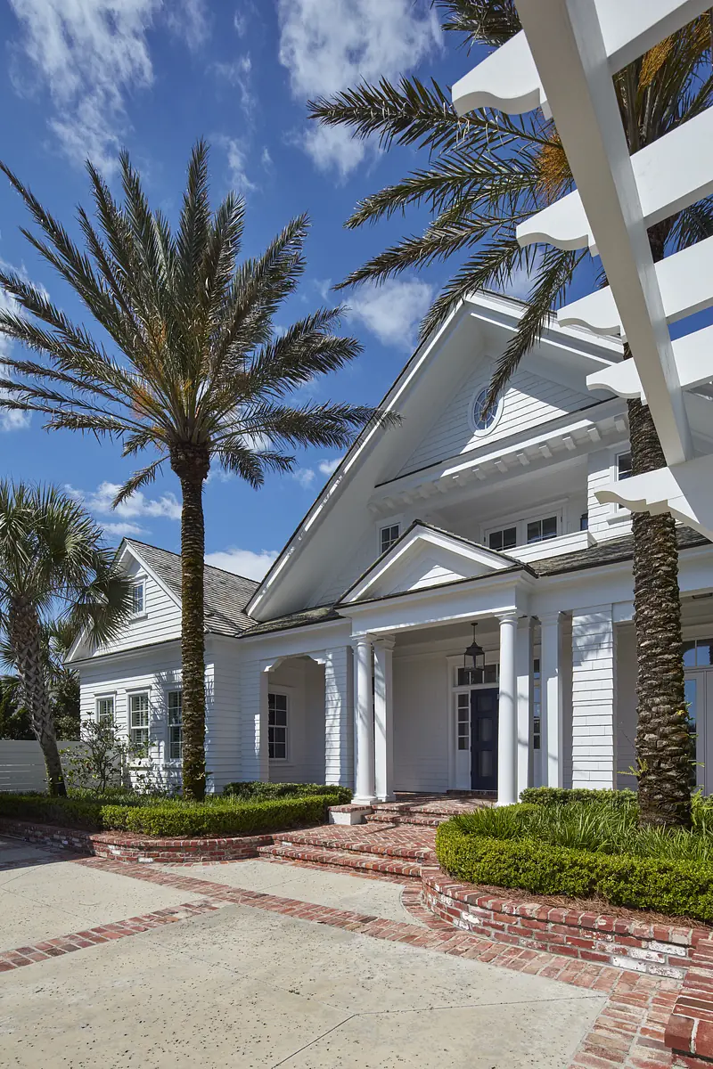 Exterior front of a house with white facade, columns, palm trees, brick steps, and decorative pergola.