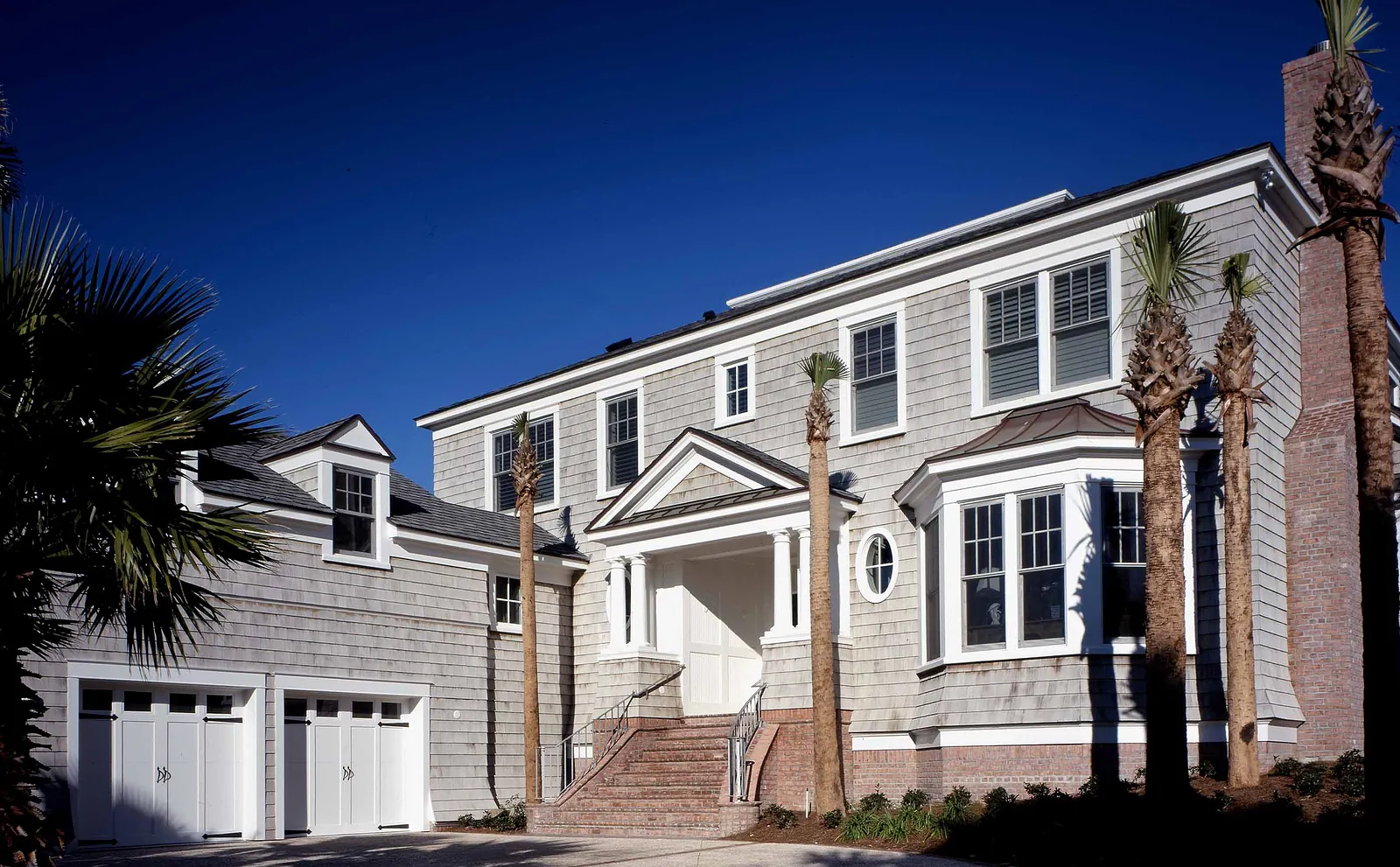 Front exterior of a house with gray siding, white trim, large windows, palm trees, and garage.