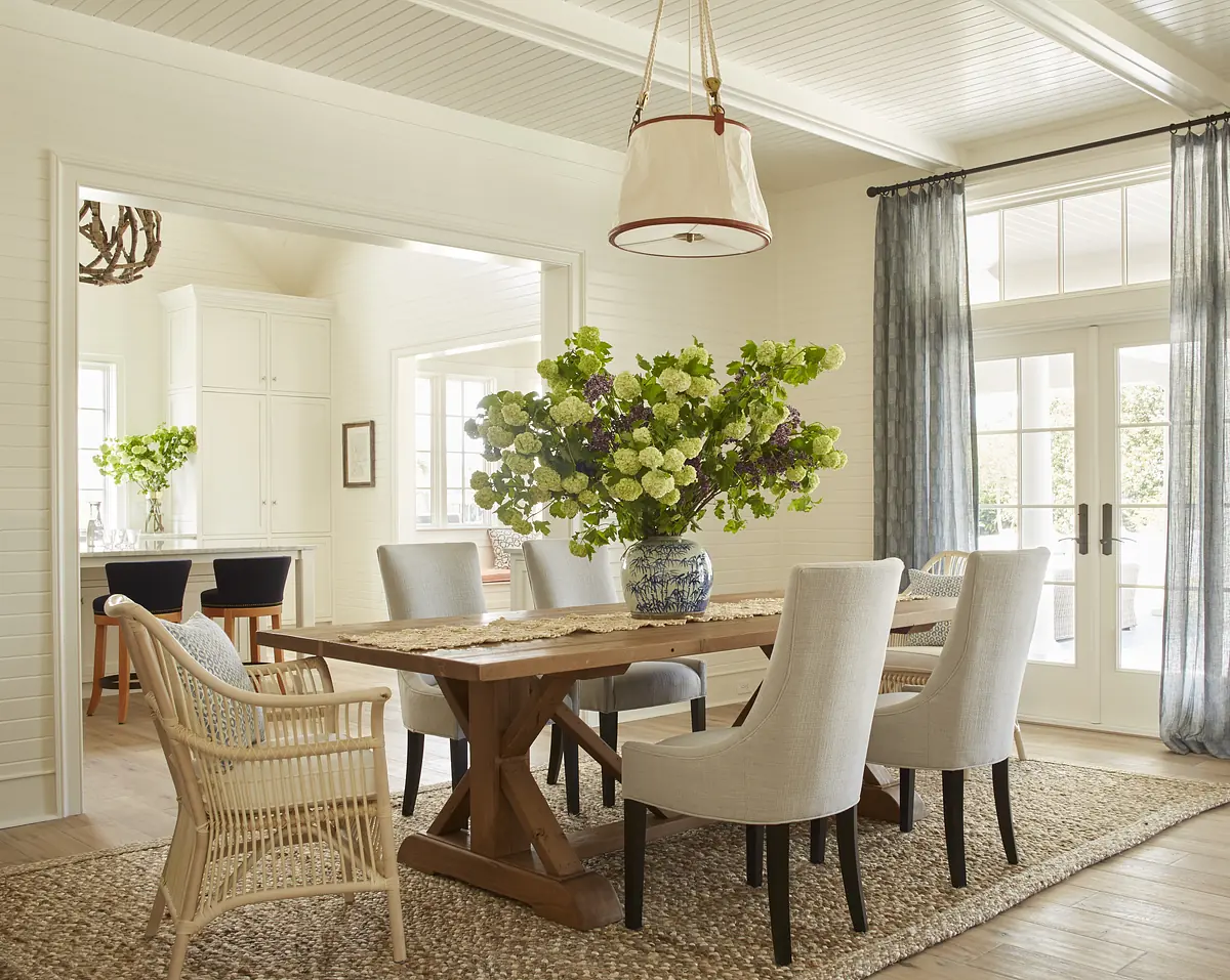 Dining room with wooden table, six chairs, floral centerpiece, woven rug, and large glass doors.