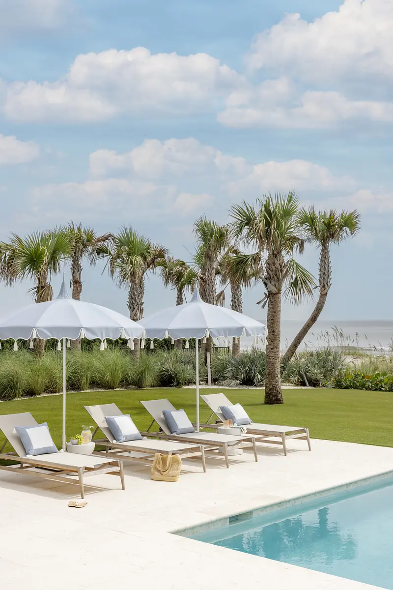 Patio area with lounge chairs, umbrellas, swimming pool, grass, and palm trees by the beach.