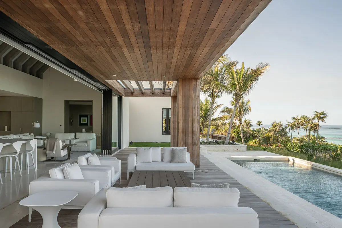 Outdoor patio with white sectional sofas, side table, wooden decking, swimming pool, and tropical plants.
