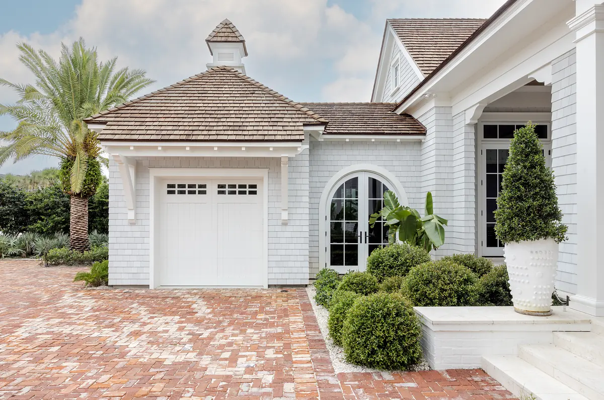 Exterior front view of a house with white facade, shingle roofing, brick driveway, and landscaping.