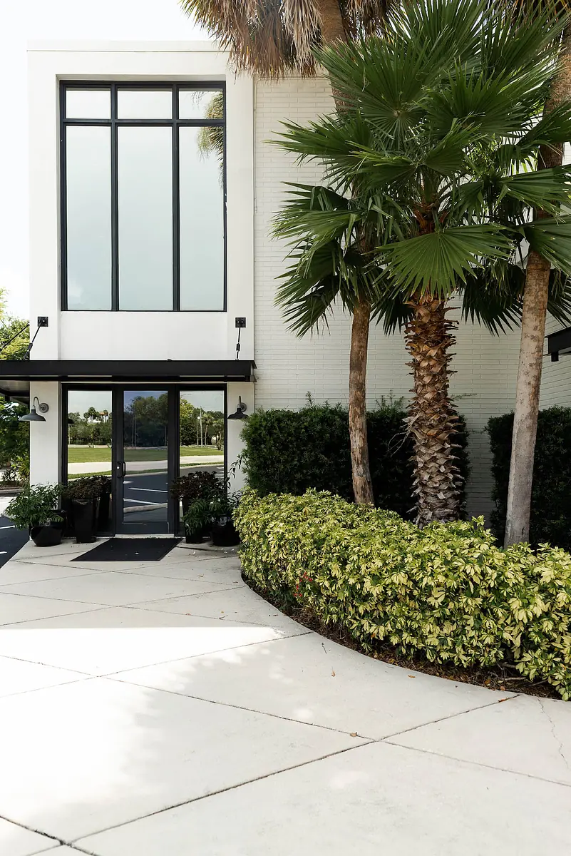 Exterior front with glass-paneled door, large windows, palm trees, and a curved concrete pathway.