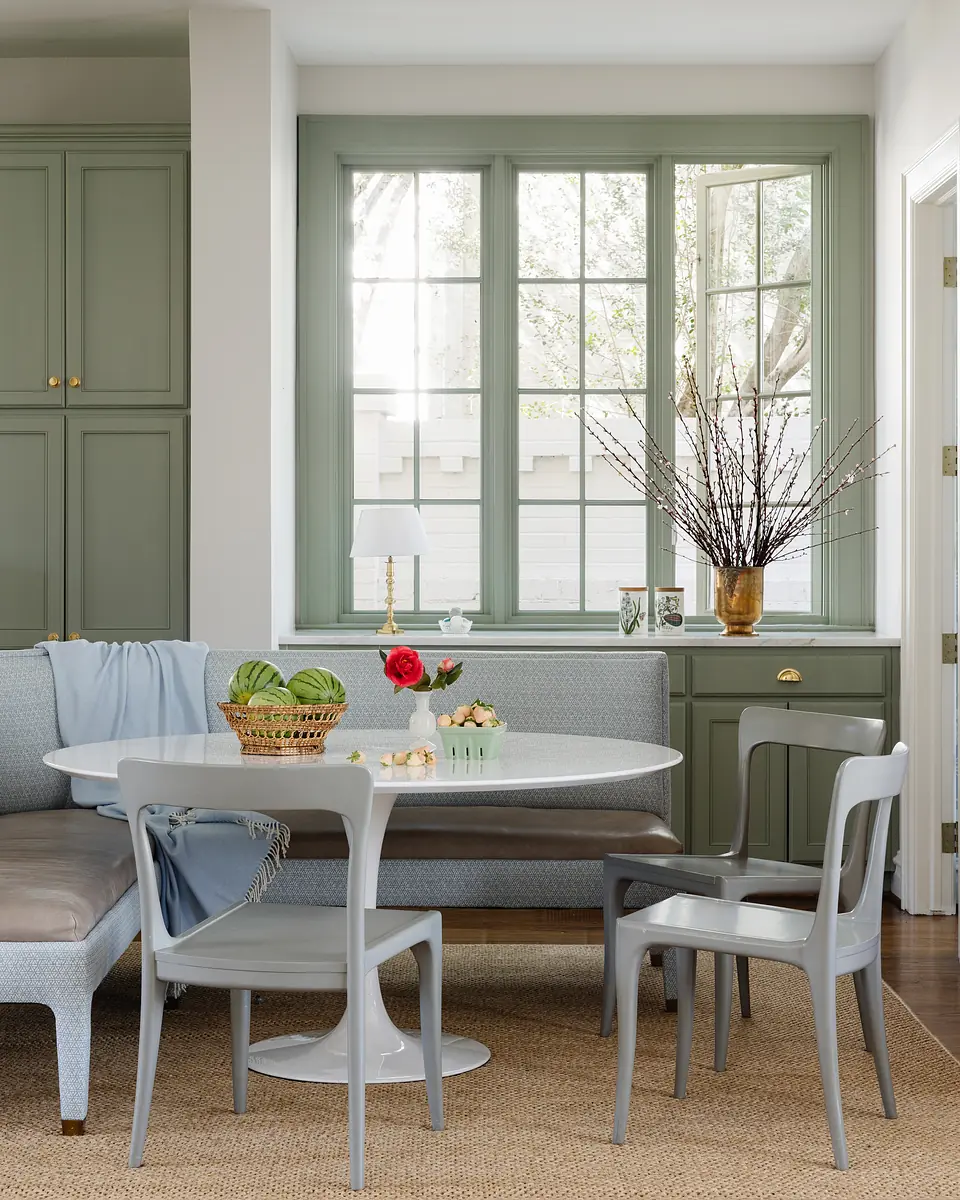 Dining room with round white table, gray chairs, light blue banquette, dark green cabinetry, and large windows.