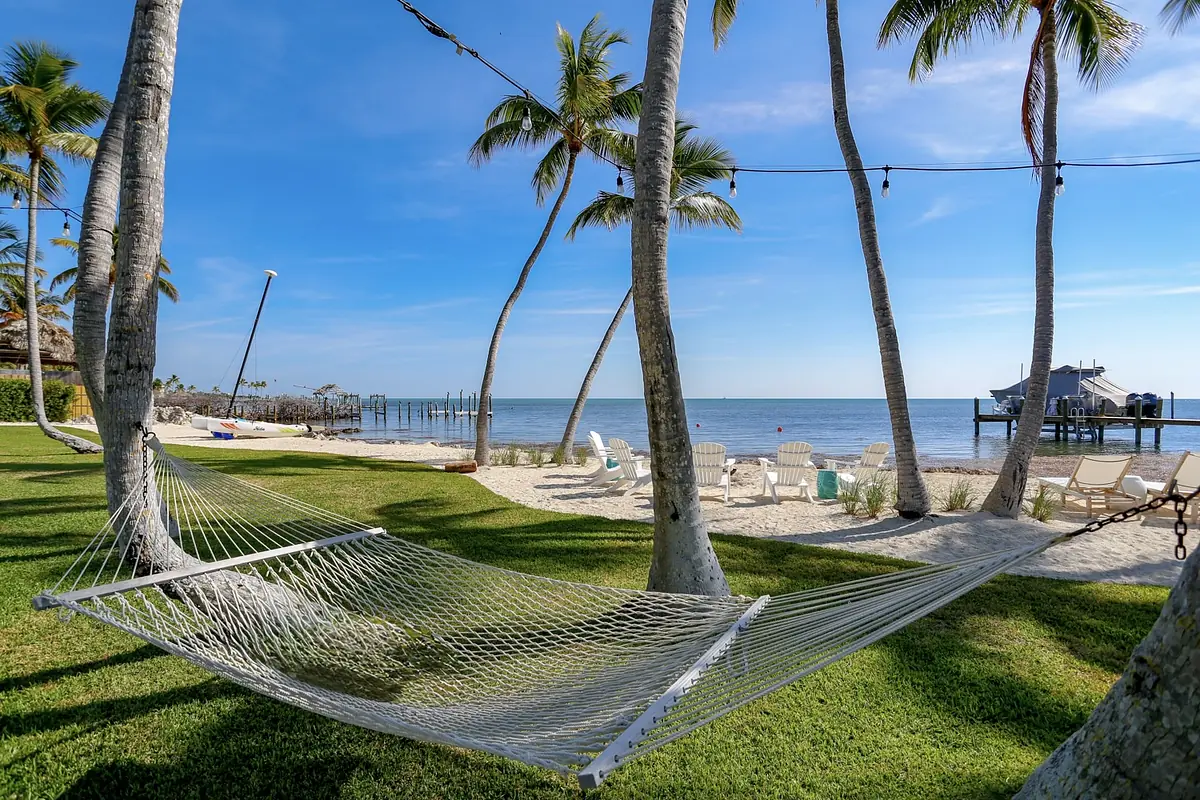 Outdoor setting with hammock, palm trees, lounge chairs, sand, and a wooden pier.