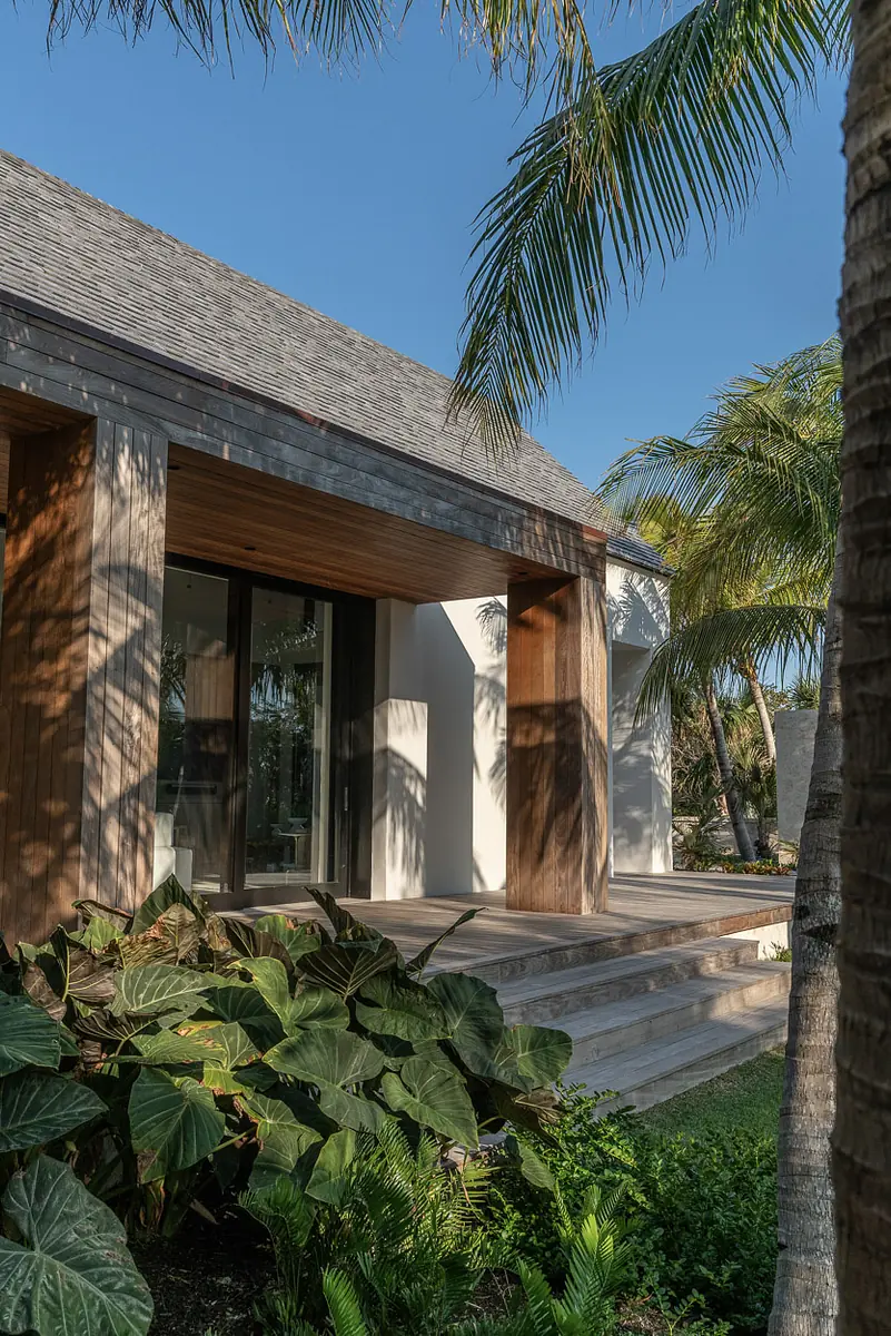 Exterior front of a house with wooden beams, large glass doors, and tropical plants.