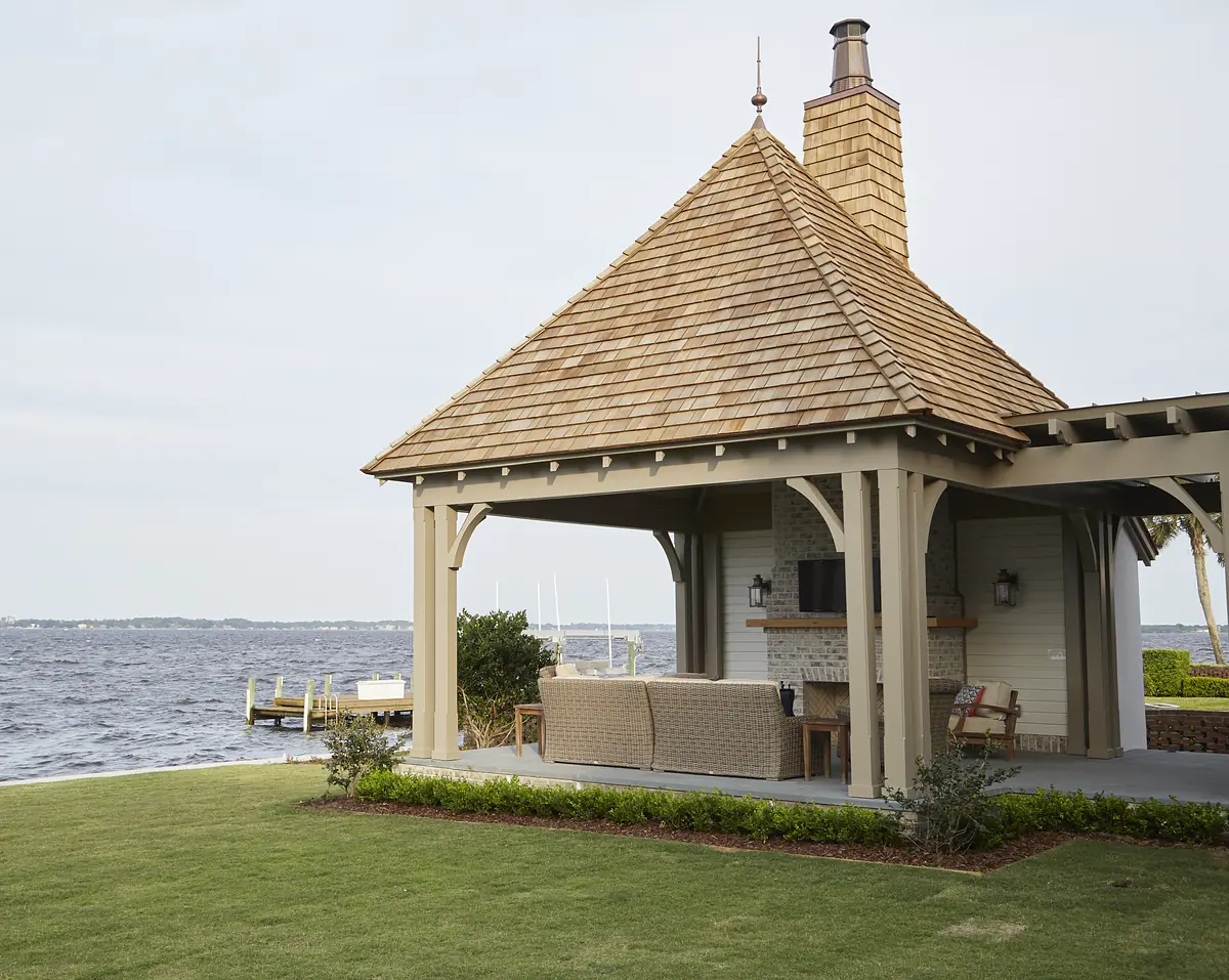 Patio with wooden structure, sectional sofa, coffee table, and television by a body of water.