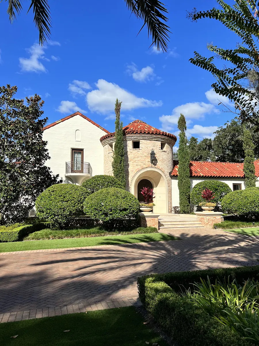Front exterior of a house with stone turret, red tile roof, wooden door, cypress trees, and paved driveway.
