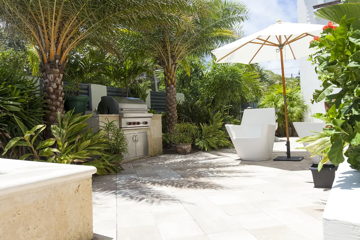 Patio with built-in grill, white chairs, large umbrella, and lush greenery.