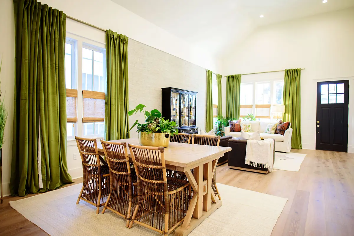 Dining room with wooden table, wicker chairs, potted plants, windows with green curtains, and a sofa.