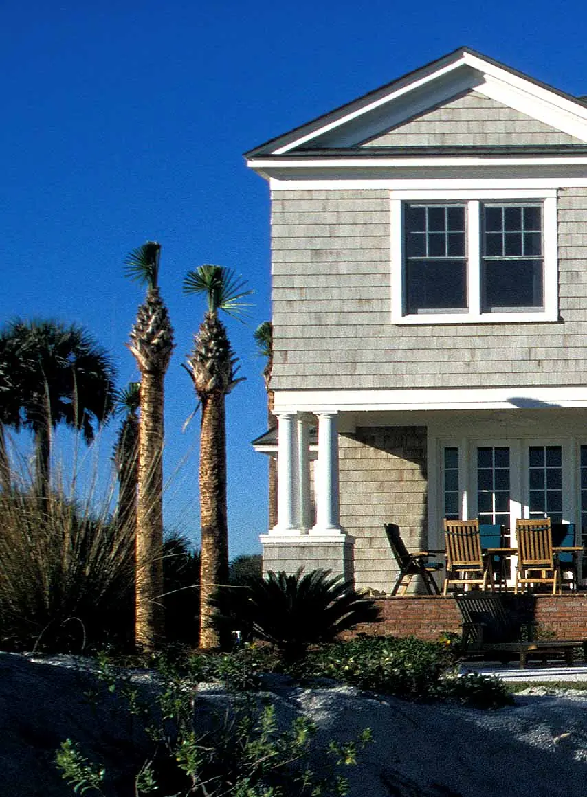 Exterior view of a house with gray shingle siding, palm trees, and a patio with wooden chairs.