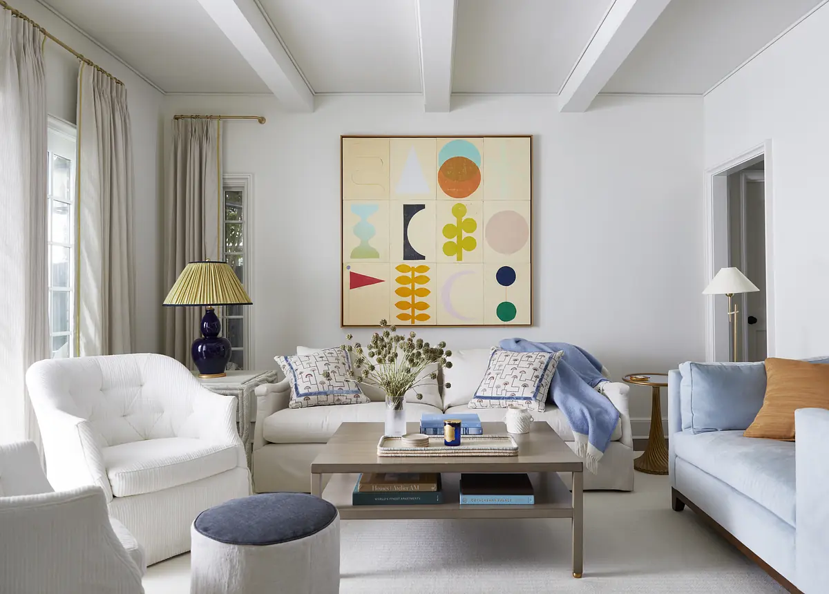 Living room with white sofas, blue throw blanket, round stool, wooden coffee table, and geometric artwork.
