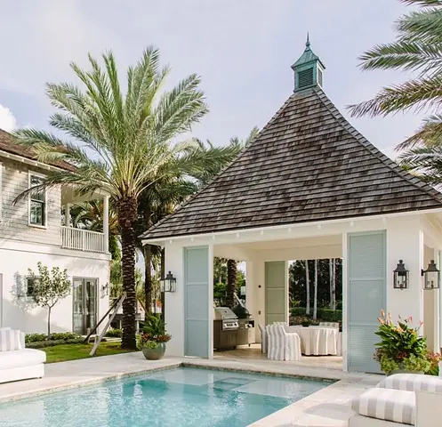 Outdoor patio with pool, built-in grill, seating area, and palm trees, featuring a peaked roof structure.