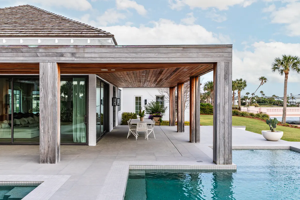 Covered patio with wooden beams, round table, chairs, potted plant, and adjacent swimming pool.