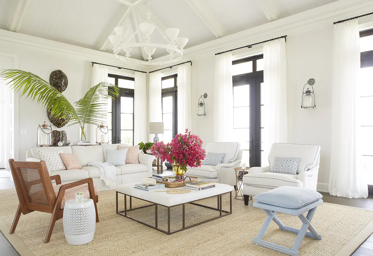 Living room with white sofa, armchairs, coffee table, area rug, plant, floral centerpiece, and tall windows with curtains