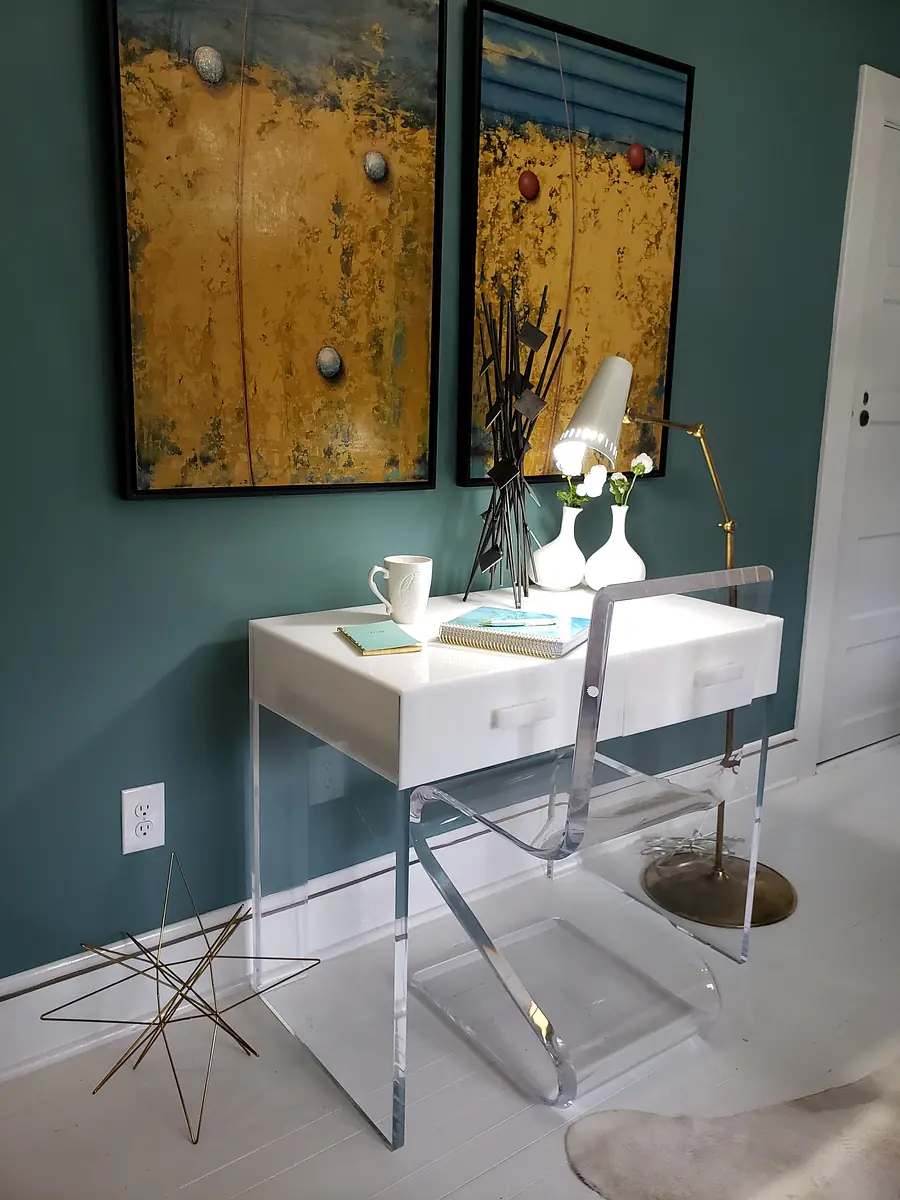 Office space with white desk, acrylic chair, gold floor lamp, paintings, and decorative objects.