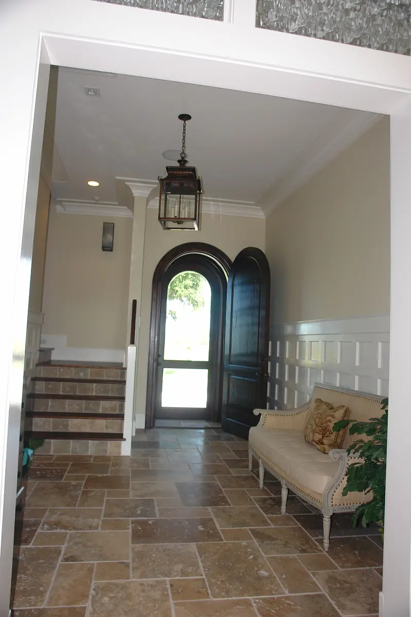 Foyer with arched wooden door, bench, staircase, stone tile floor, and decorative wall paneling.