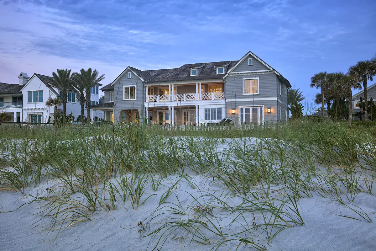 Beach house with light gray exterior, balconies, large windows, sandy terrain, and palm trees.