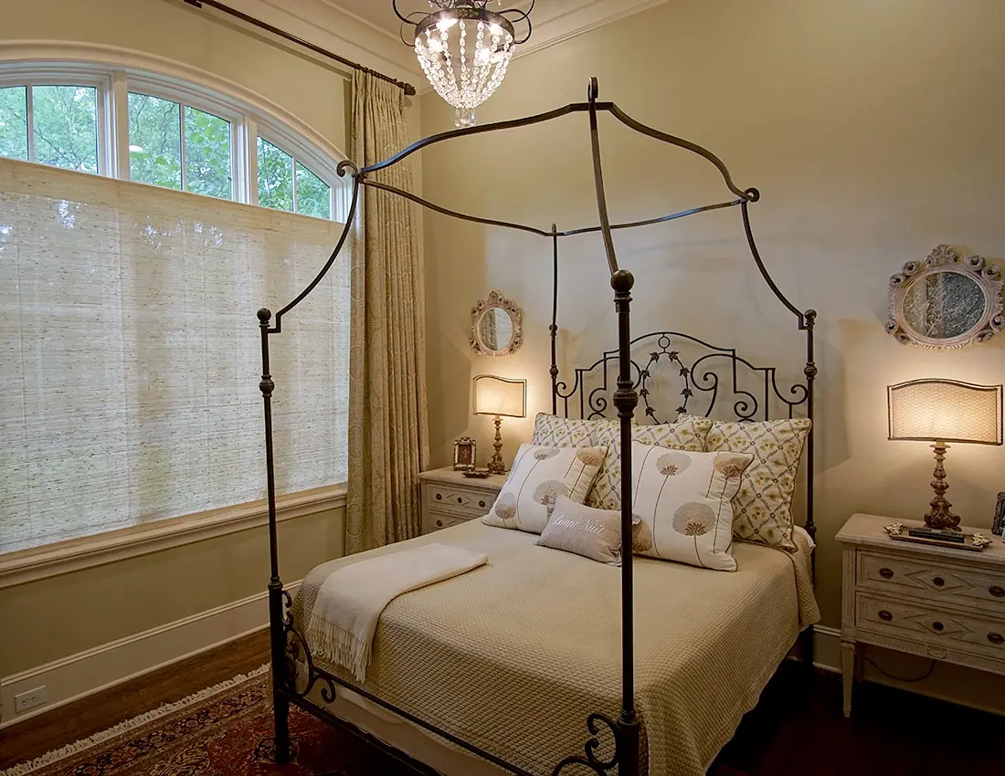Bedroom with metal canopy bed, beige quilt, nightstands, lamps, and large windows with blinds