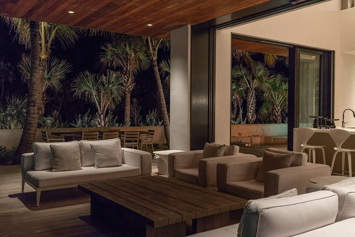 Patio with white outdoor sofas, wooden coffee table, and dining set amidst palm trees.