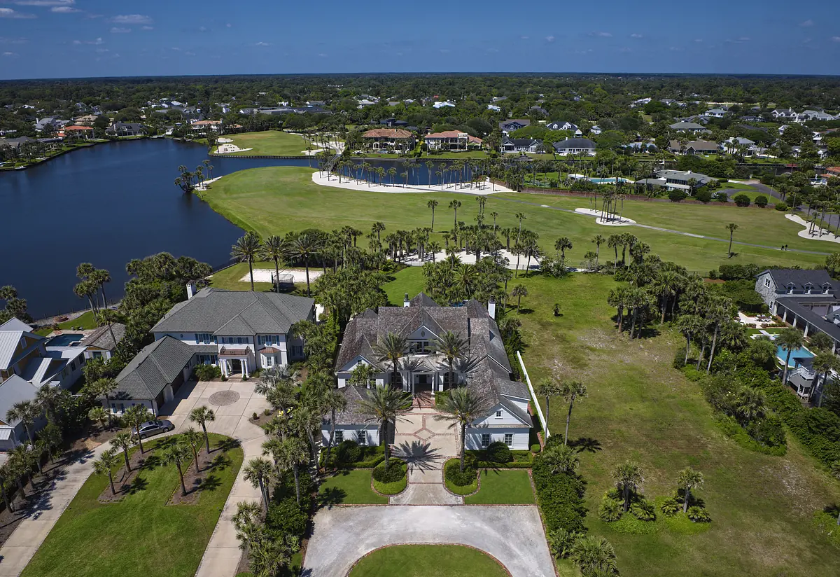 Aerial view of a property with gray shingle roof, landscaped lawns, palm trees, lake, and golf course.