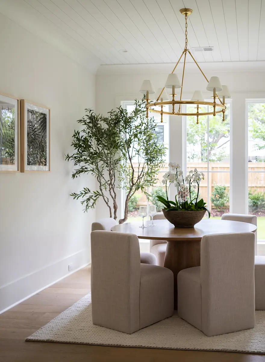 Dining room with round wooden table, upholstered chairs, potted plant, windows, artworks, and chandelier