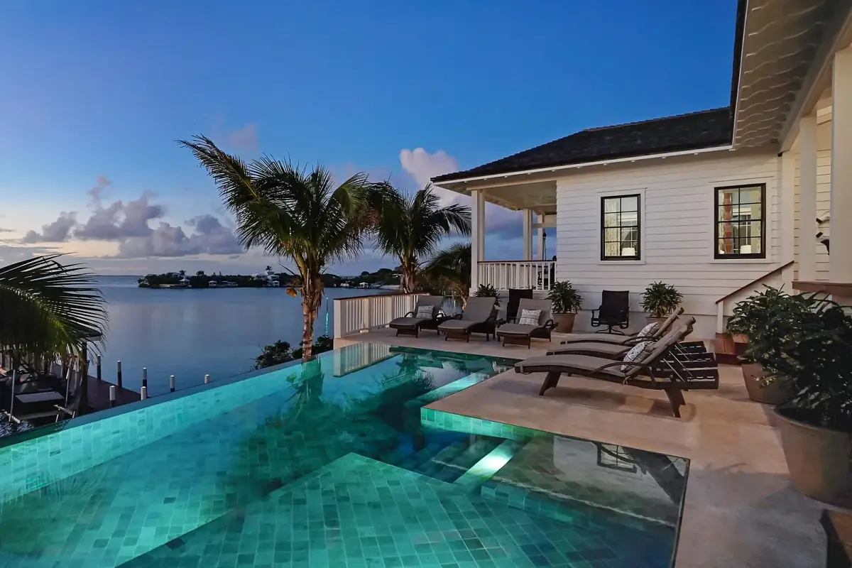 Exterior rear with swimming pool, sun loungers, palm trees, water view, and house with white siding at twilight.