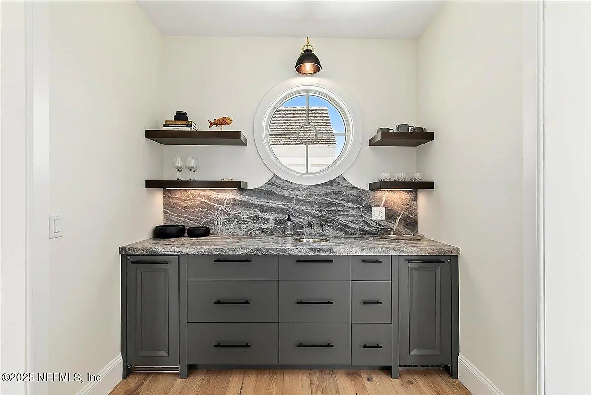 Butler's pantry with gray cabinets, marble countertop, circular window, and glassware on floating shelves.