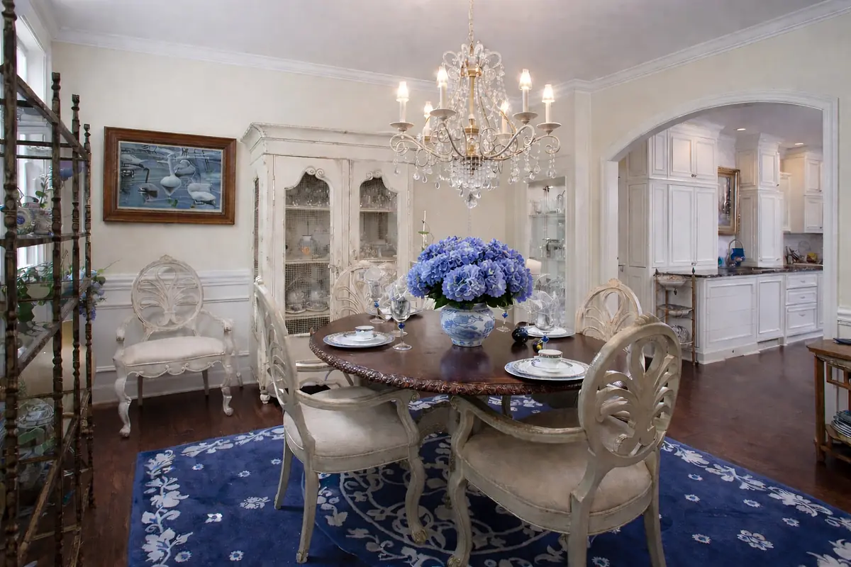 Dining room with round wooden table, floral arrangement, upholstered chairs, glass-front cabinet, and chandelier.