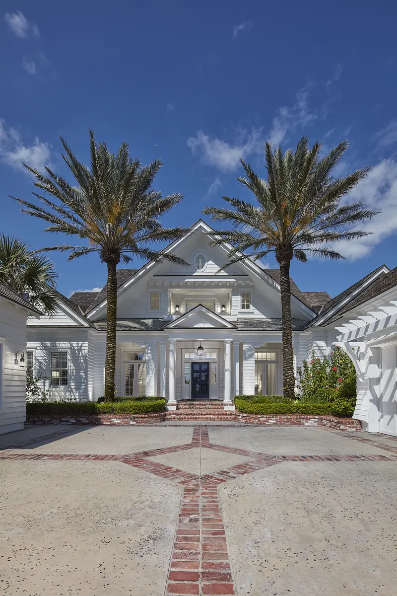 Front exterior of a house with palm trees, brick pathway, double doors, and gabled roof.