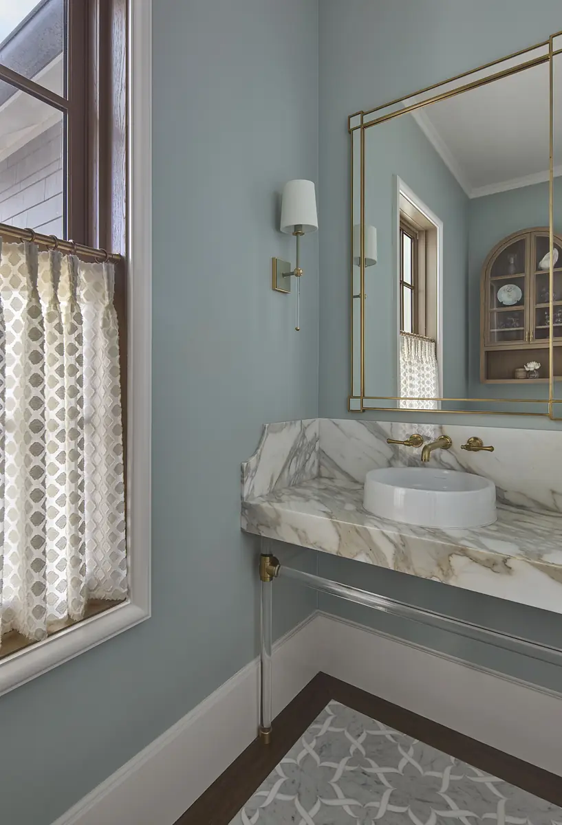 Powder room with marble sink, brass faucet, large mirror, patterned curtains, and decorative rug.