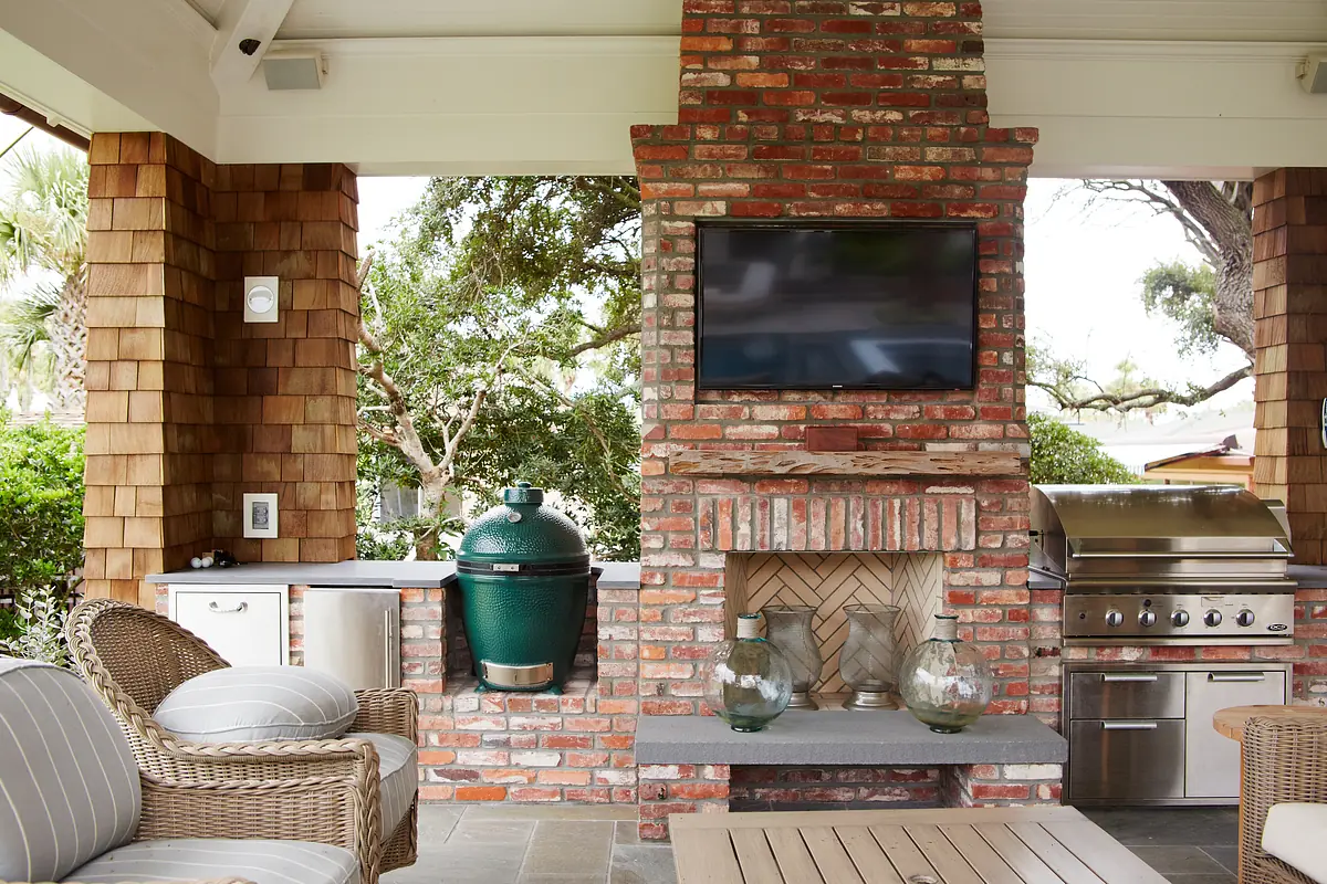 Patio with brick fireplace, outdoor kitchen, wicker chairs, and mounted television surrounded by greenery.