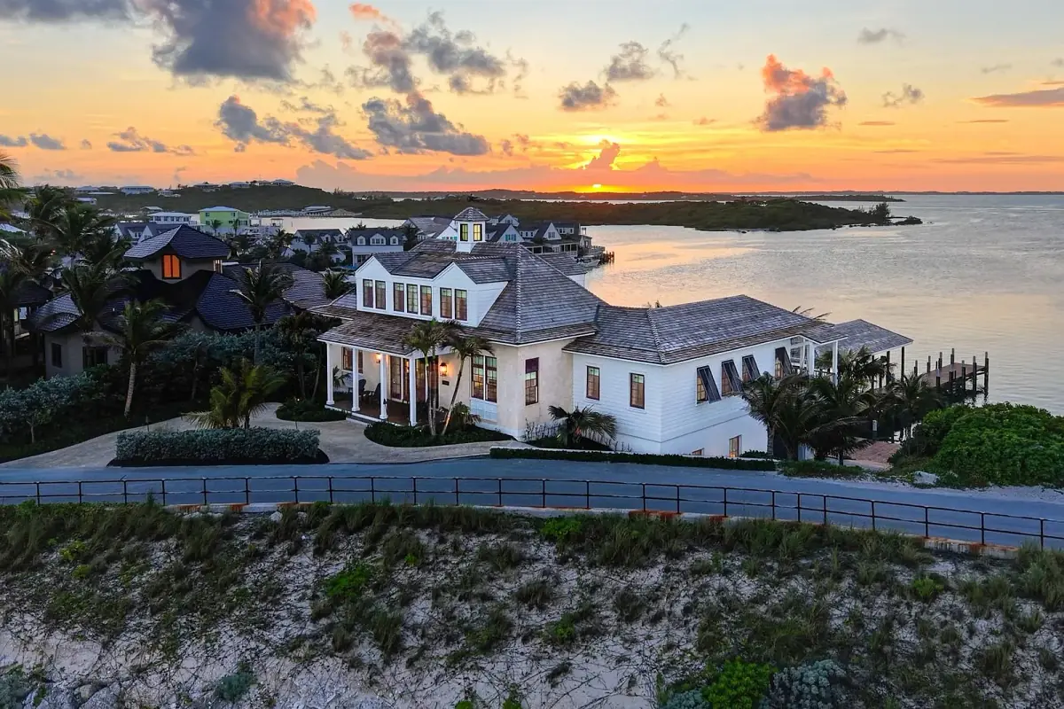 Coastal home with white exterior, multiple gables, wraparound porch, palm trees, and a sunset view over water.
