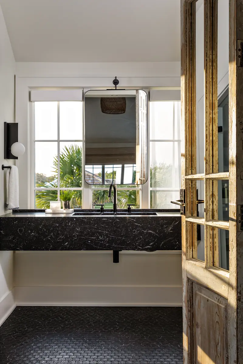 Bathroom with black marble countertop, large mirror, pebble tile floor, and wooden door
