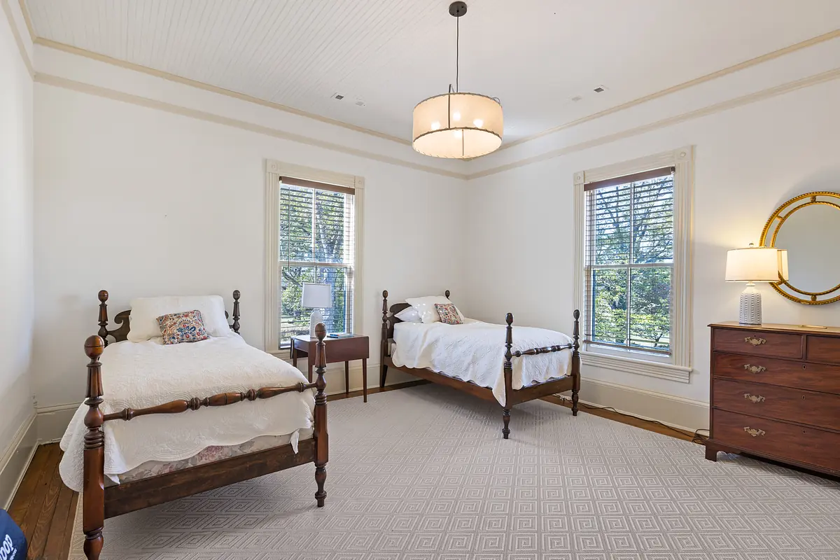 Bedroom with two wooden twin beds, nightstand, lamp, dresser, and round mirror with natural light from windows.